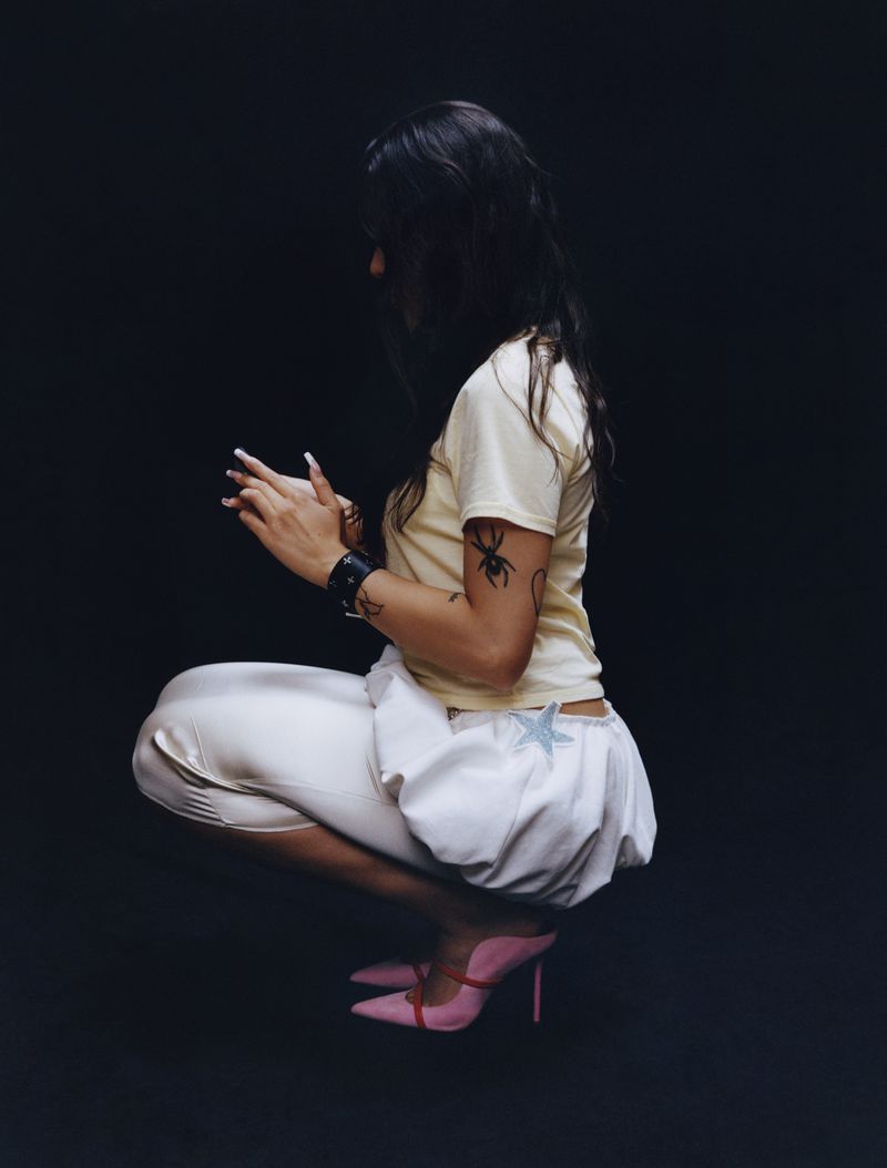 Side view of a woman in a cream t-shirt, white shorts and pink heels, squatting in front of a dark background, showcasing an alternative style.