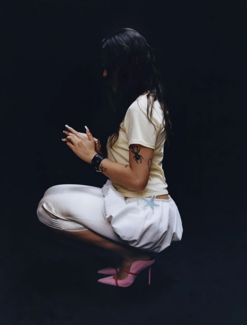 Side view of a woman in a cream t-shirt, white shorts and pink heels, squatting in front of a dark background, showcasing an alternative style.