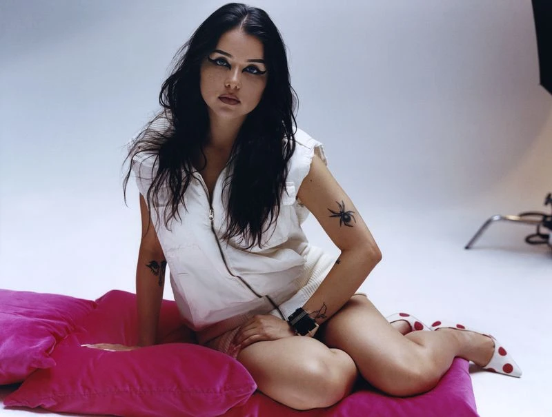 A person sits on a pink pillow in a studio, wearing a white vest, tan shorts, black bracelets, and polka-dot shoes, eyes looking at camera.