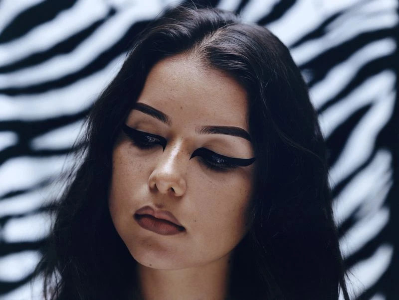 Close-up of a woman with black hair, wearing heavy black eyeliner and neutral lipstick, posed against a white and black zebra-print backdrop.