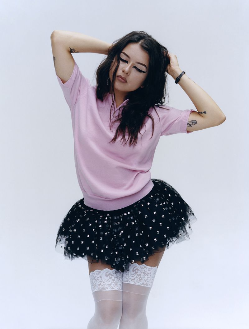 Young woman in pink top and black tutu poses in a white studio, her hands behind her head. She is looking down.