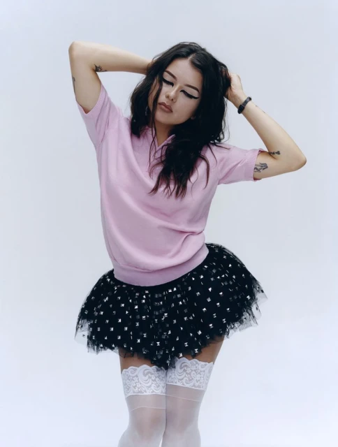 Young woman in pink top and black tutu poses in a white studio, her hands behind her head. She is looking down.