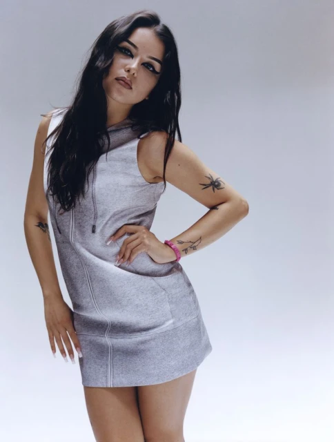 Portrait of a young woman with long, dark hair wearing a gray dress with tattoos on her arms in a studio setting against a white backdrop.