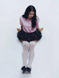 Young woman in a pink polo shirt and black tutu skirt sitting on a white seamless background with white thigh-high stockings and heels.