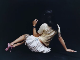 Woman with dark hair seated on black seamless floor wearing yellow t-shirt, white skirt, and pink high heels.