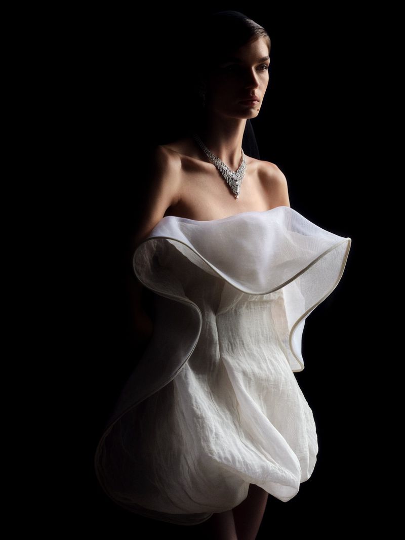 Elegant woman in a white dress and diamond necklace poses in a dramatic fashion photo against a dark backdrop, showcasing beauty and style.