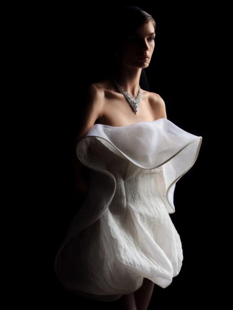 Elegant woman in a white dress and diamond necklace poses in a dramatic fashion photo against a dark backdrop, showcasing beauty and style.