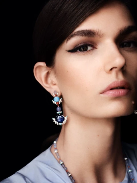 Close-up of a woman with dramatic eye makeup wearing elegant gemstone earrings, captured against a black background in a portrait.