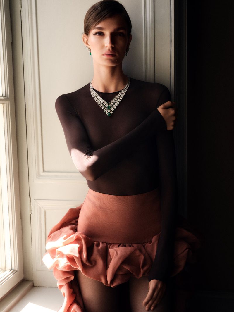 An adult female model in a brown bodysuit and pink ruffle skirt poses next to a white door wearing jewelry in a studio-lit fashion shot.
