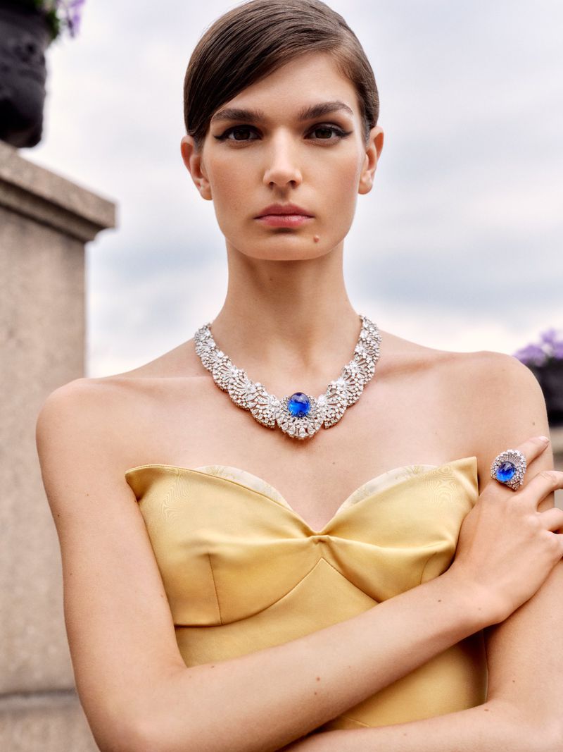 Woman in a yellow dress and diamond necklace and ring with a blue gem, outdoors. Shot at eye-level, with shallow depth-of-field.