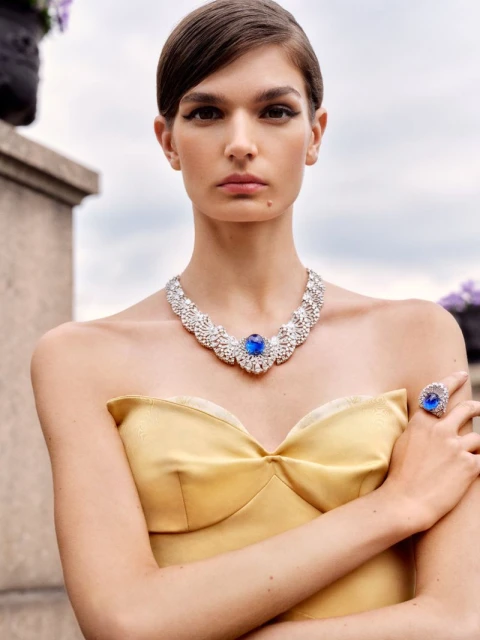 Woman in a yellow dress and diamond necklace and ring with a blue gem, outdoors. Shot at eye-level, with shallow depth-of-field.