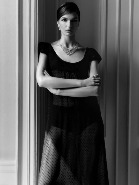 Portrait of a woman in a black dress and necklace standing in front of a white doorway, captured in a black and white photograph.