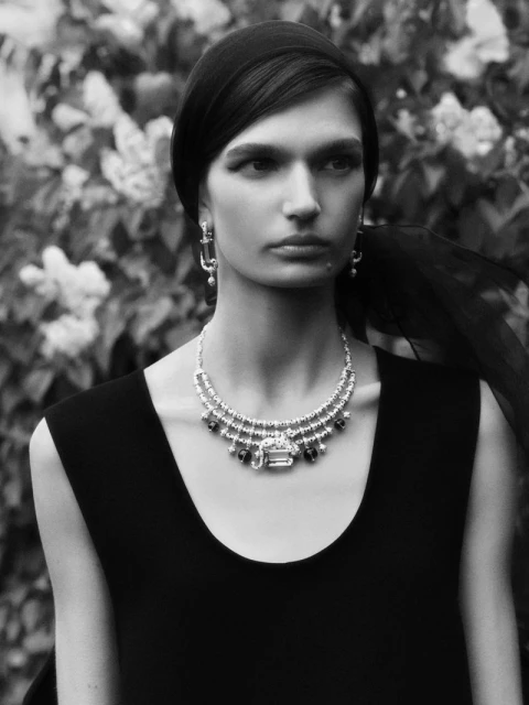 Black and white portrait of a woman with headscarf wearing a statement jewel necklace and earrings standing in front of a floral bush.