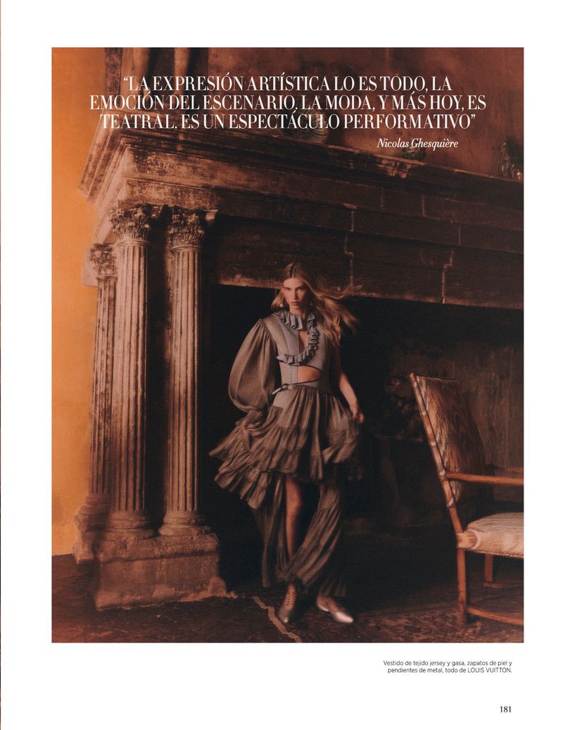 Woman in a ruffled brown dress poses in front of a fireplace with columns indoors, captured in a fashion editorial photograph.