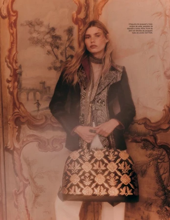 Editorial fashion shot of a woman in a blazer holding a floral embroidered handbag, posing in front of a painted landscape.
