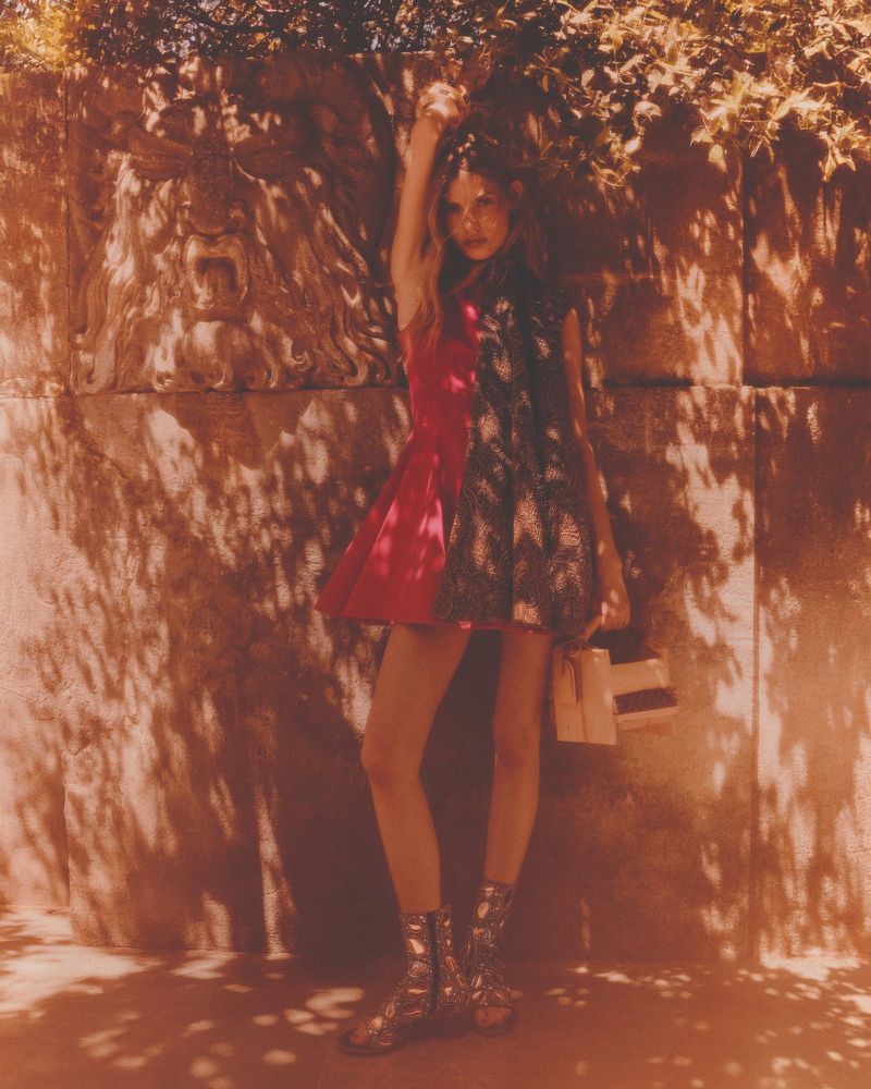 A young adult woman poses outdoors in a red and black dress with short metallic boots, holding a shopping bag in diffused sunlight.