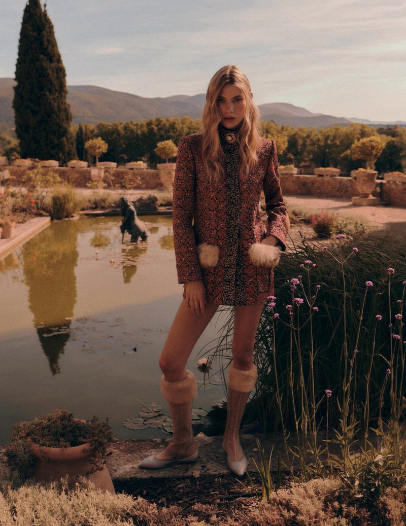 A woman in a patterned coat poses in a garden setting with a pond reflecting the sky and surrounding greenery, capturing a blend of fashion and nature.