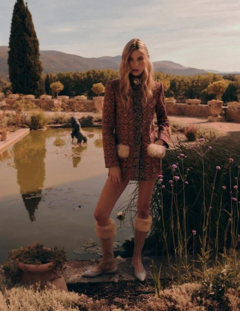 A woman in a patterned coat poses in a garden setting with a pond reflecting the sky and surrounding greenery, capturing a blend of fashion and nature.