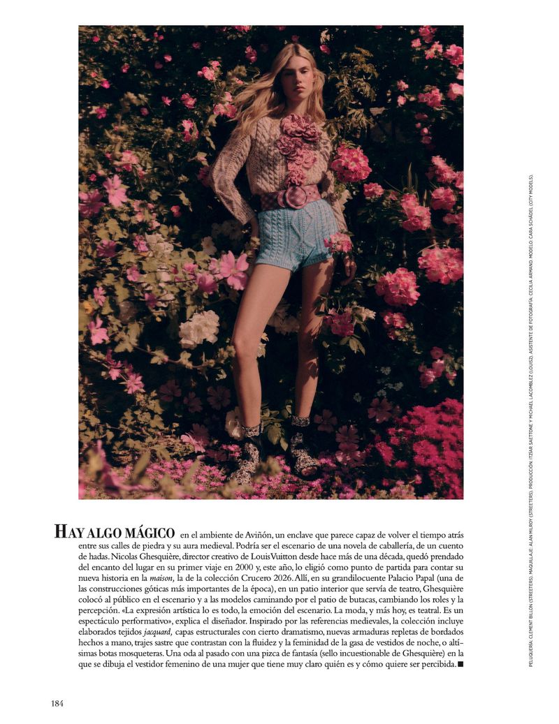 A woman poses in a garden wearing light-colored knitwear and blue shorts, surrounded by pink flowers, captured with a soft, diffused light.