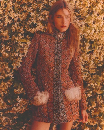 A woman in a decorative dress poses in front of a wall of small white flowers, bathed in soft lighting with a shallow depth of field.