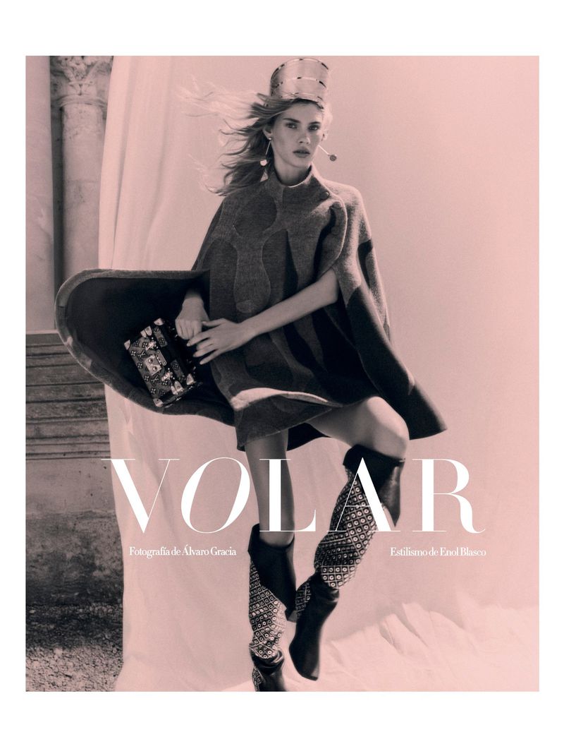Fashion editorial shot of a blond woman wearing a crown and boots with a patterned knit cape and holding a small purse.
