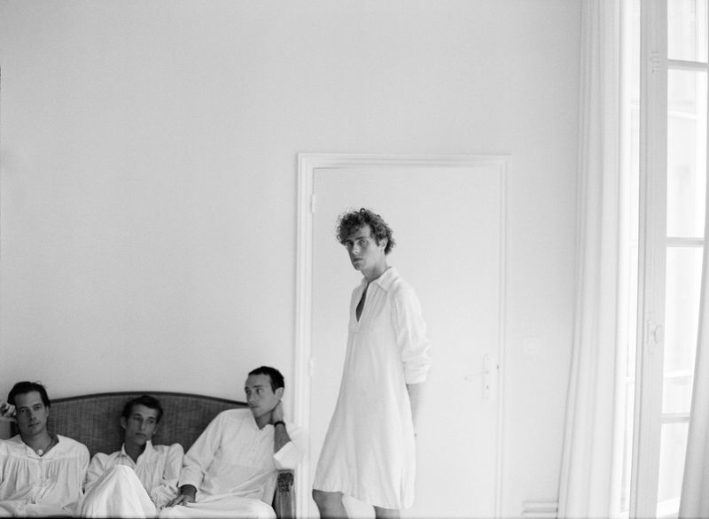Black and white photo of four young adult males in white shirts, one standing near a door, the others seated on a rattan couch.