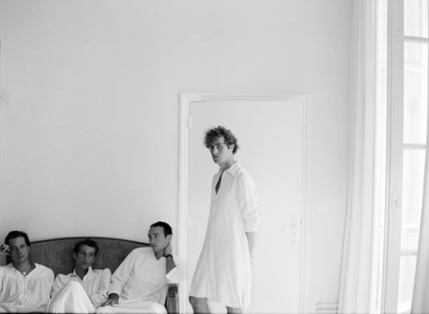 Black and white photo of four young adult males in white shirts, one standing near a door, the others seated on a rattan couch.