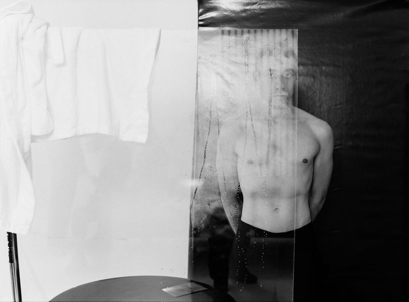 Monochrome image featuring an adult man partially obscured behind glass next to a suspended white shirt against a contrasting background.