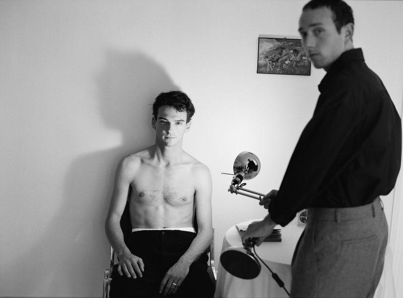 Black and white shot of two young adult men in a room with a lamp and painting on the wall, indoor setting, medium shot.
