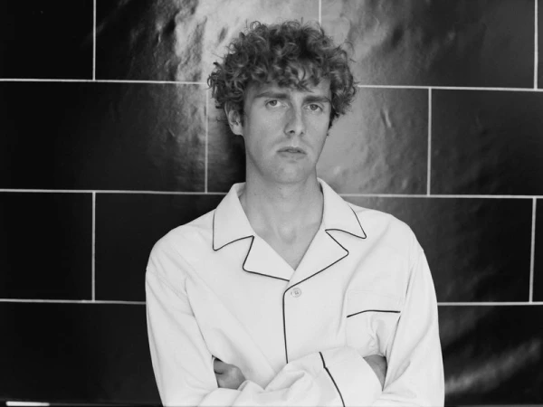 Portrait of a young adult man with curly hair, wearing a white shirt and standing against a glossy dark tile wall, arms crossed.