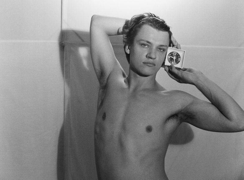 A shirtless young adult male with short hair holds a square alarm clock next to his face in a brightly lit indoor space.