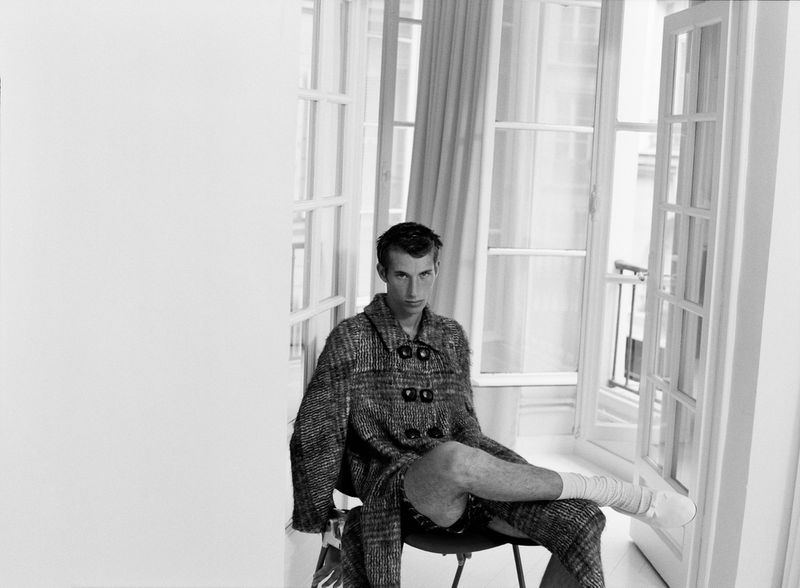 Black and white studio portrait of a young adult male seated in a chair, wearing a textured overcoat near window light.