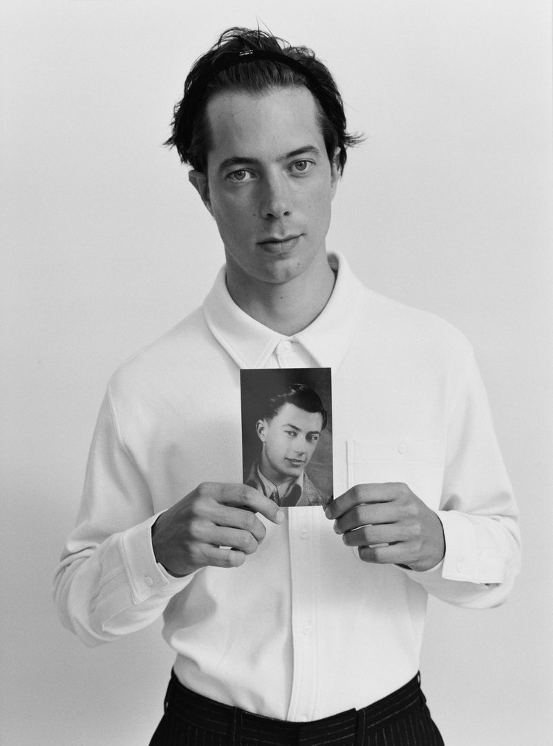 Monochrome portrait of a young man holding a vintage photo of another man against a plain background, wearing a white collared shirt.