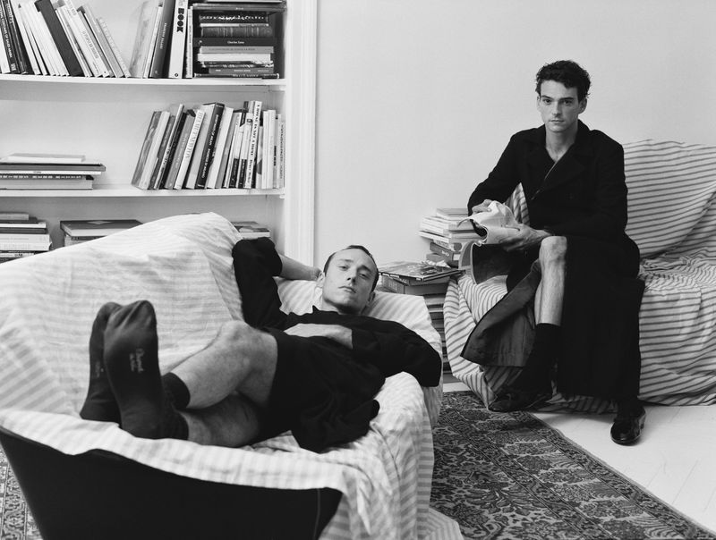 Black and white portrait of two adult men in a domestic interior, one reclining on a couch, the other sitting holding clothing.