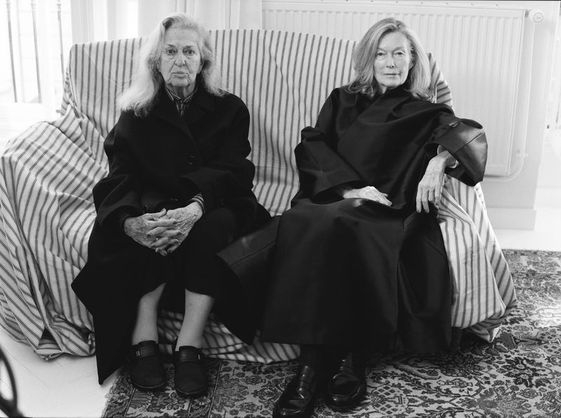 Black-and-white photograph of two mature women sitting on a striped couch, posing for the camera indoors in a natural lighting setting.