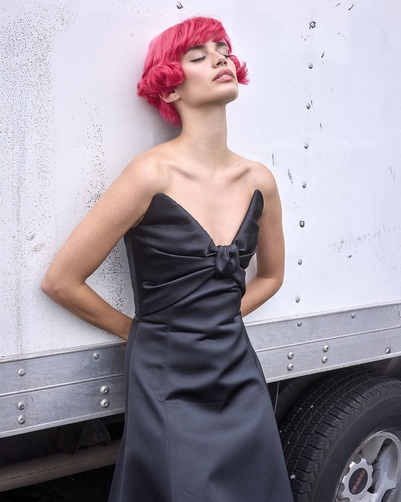 A woman with pink hair wears a black strapless dress in an outdoor fashion portrait, leaning against the side of a white truck.