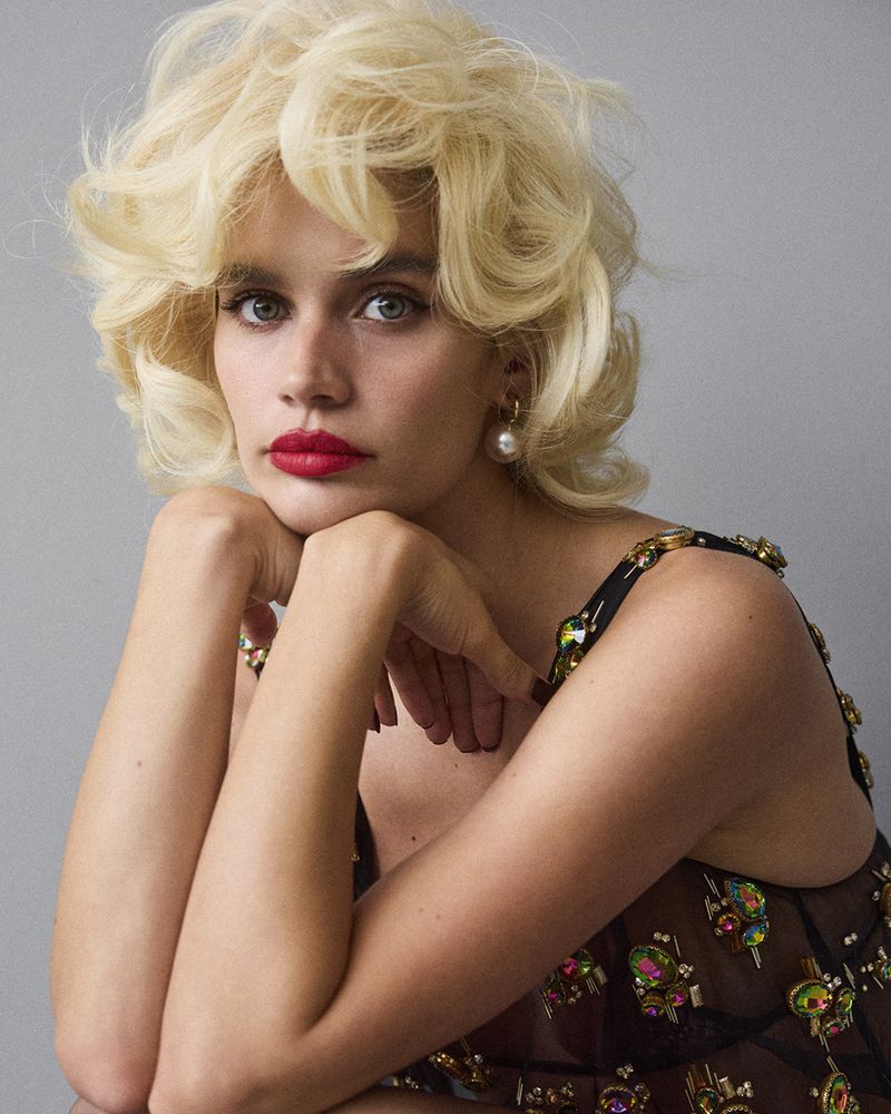 Close-up of a blonde woman with red lipstick, pearl earrings and a semi-sheer beaded top posing indoors with her arms supporting her chin.