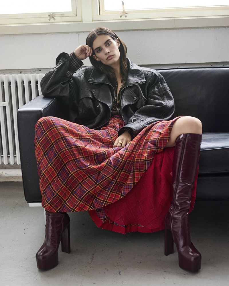 A woman sits on a black couch wearing a black jacket, a red and yellow plaid skirt, and knee-high burgundy platform boots in an indoor setting.