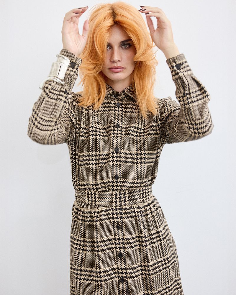 A woman with orange layered hair wears a long-sleeved plaid dress and silver bracelet in a brightly lit, full shot against white.
