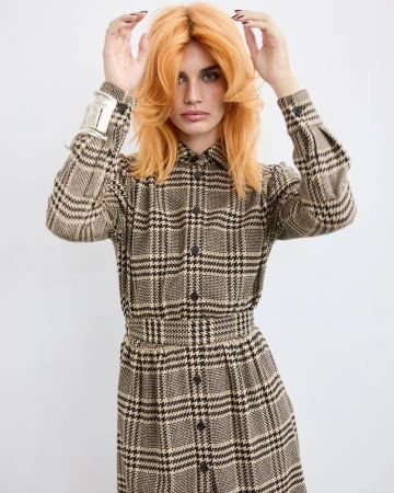 A woman with orange layered hair wears a long-sleeved plaid dress and silver bracelet in a brightly lit, full shot against white.