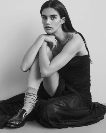 Black and white portrait of a young woman sitting on the floor, wearing a dark dress, socks and loafers, with a serene facial expression.