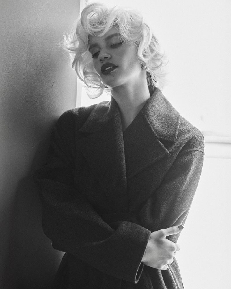 Model Sara Sampaio with blonde vintage-style curls wearing a dark coat in a black and white portrait for Behind the Blinds.