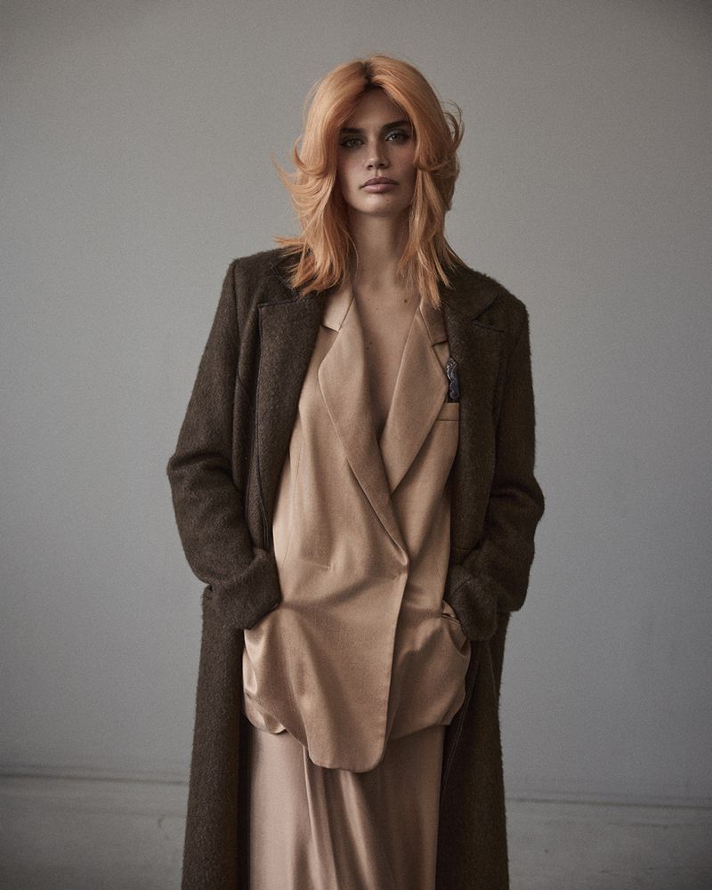 A solo female adult poses in a long brown coat and tan outfit against a neutral gray background in a studio shot with medium framing.
