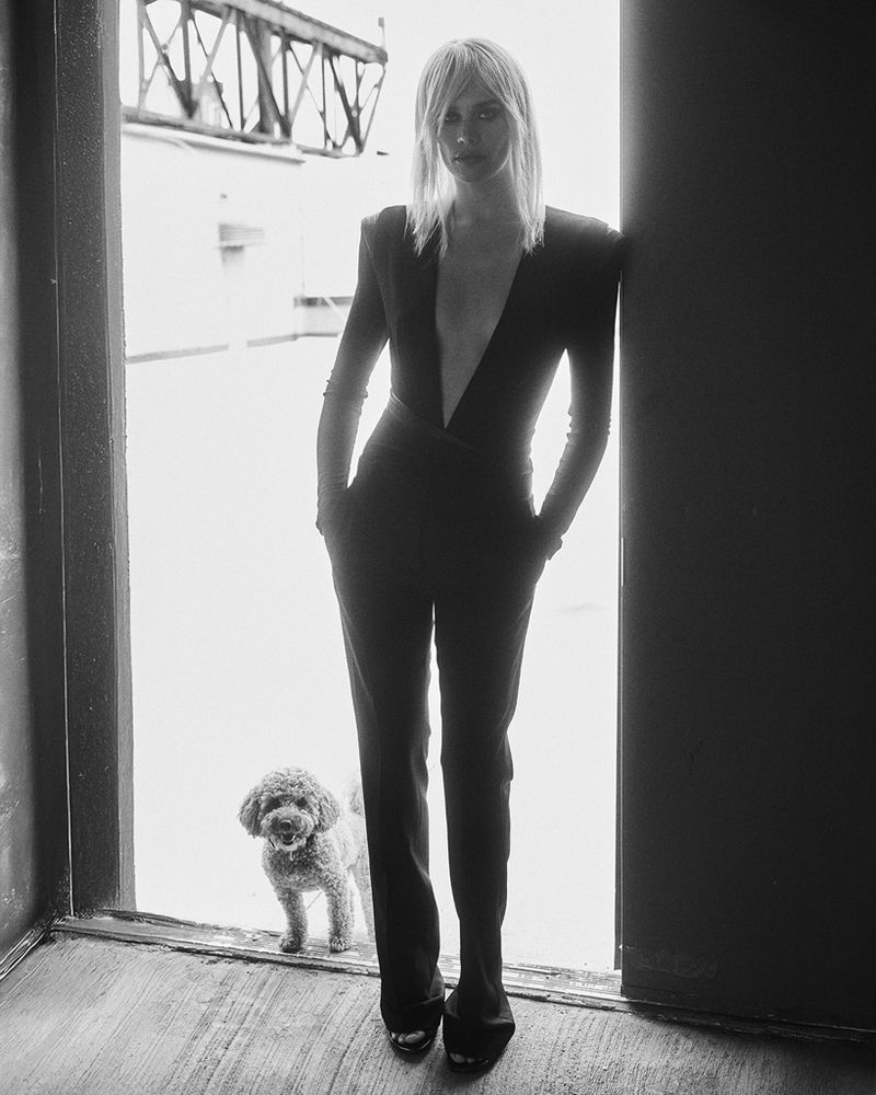 Black and white studio shot of a woman in a black jumpsuit with a plunging neckline, accompanied by a poodle near an open doorway.