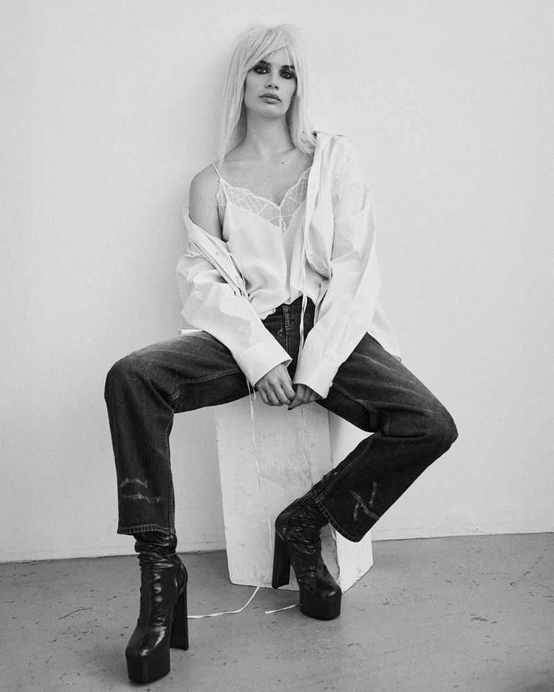 A woman with platinum blonde hair poses in a studio wearing a white top, blue jeans, and platform boots against a white background.