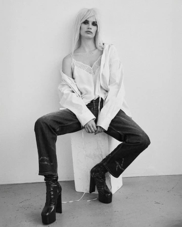 A woman with platinum blonde hair poses in a studio wearing a white top, blue jeans, and platform boots against a white background.