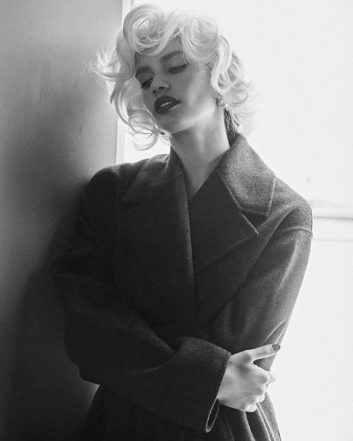Model Sara Sampaio with blonde vintage-style curls wearing a dark coat in a black and white portrait for Behind the Blinds.