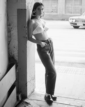 Model Sara Sampaio wearing a light bra top and dark jeans leaning against a concrete pillar in a black and white editorial image.