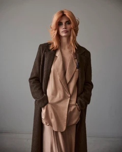 Model Sara Sampaio with orange hair wearing a brown coat and beige silk suit against a grey studio wall.