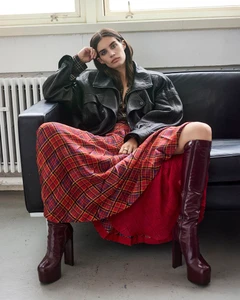 Model Sara Sampaio seated on a black leather sofa wearing an oversized black leather jacket and a red plaid skirt